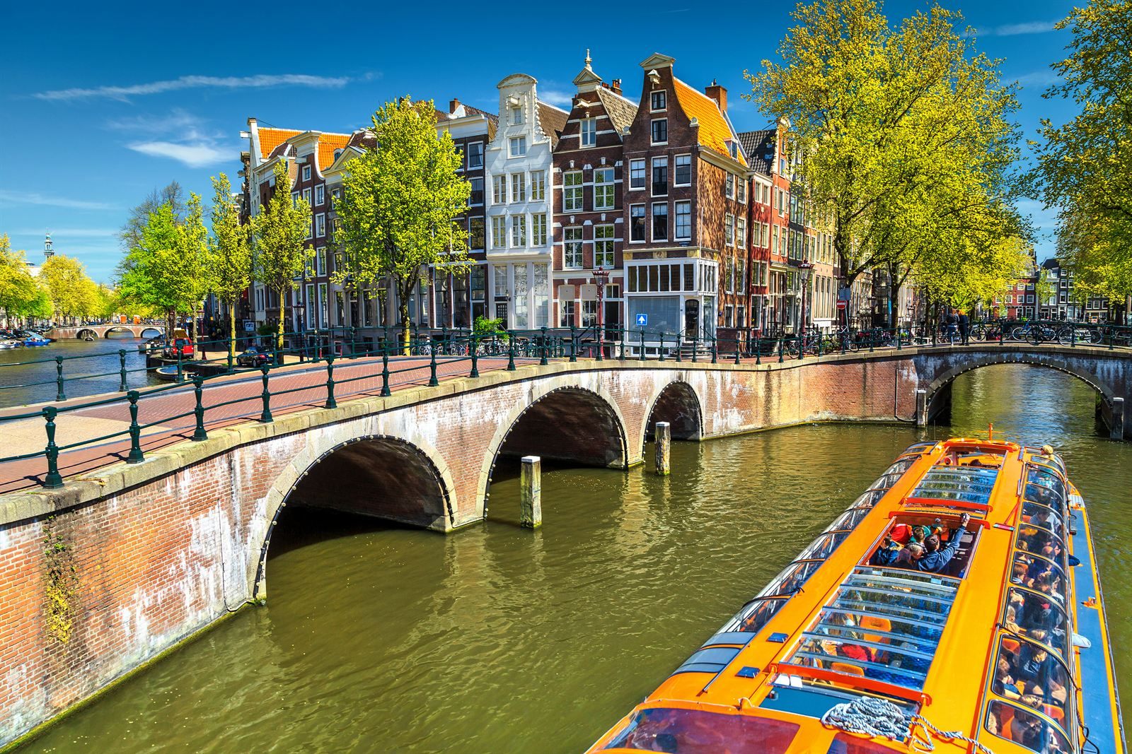 Glass-roofed canal boat passing under an arched brick bridge lined with Amsterdam gabled houses