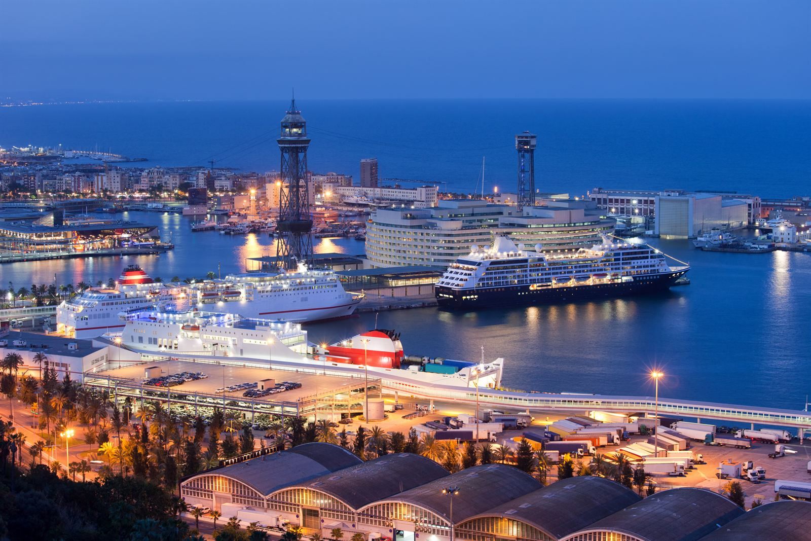 Cruise ships berthed in Barcelona harbor at dusk, with the cable car tower and city lights behind