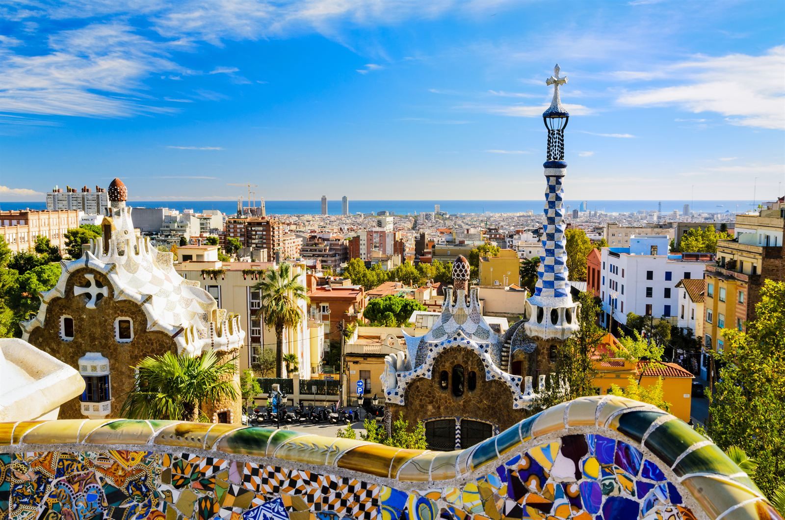 Mosaic-tiled bench and gingerbread-style gatehouses at Park Güell, with Barcelona and the Mediterranean beyond
