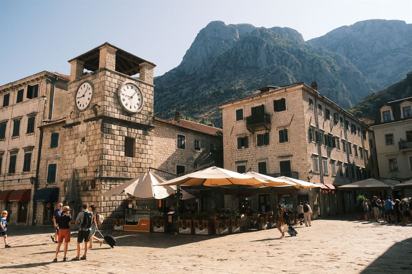 Clock tower and café umbrellas on the main square inside Kotor&rsquo;s old town, with mountains behind