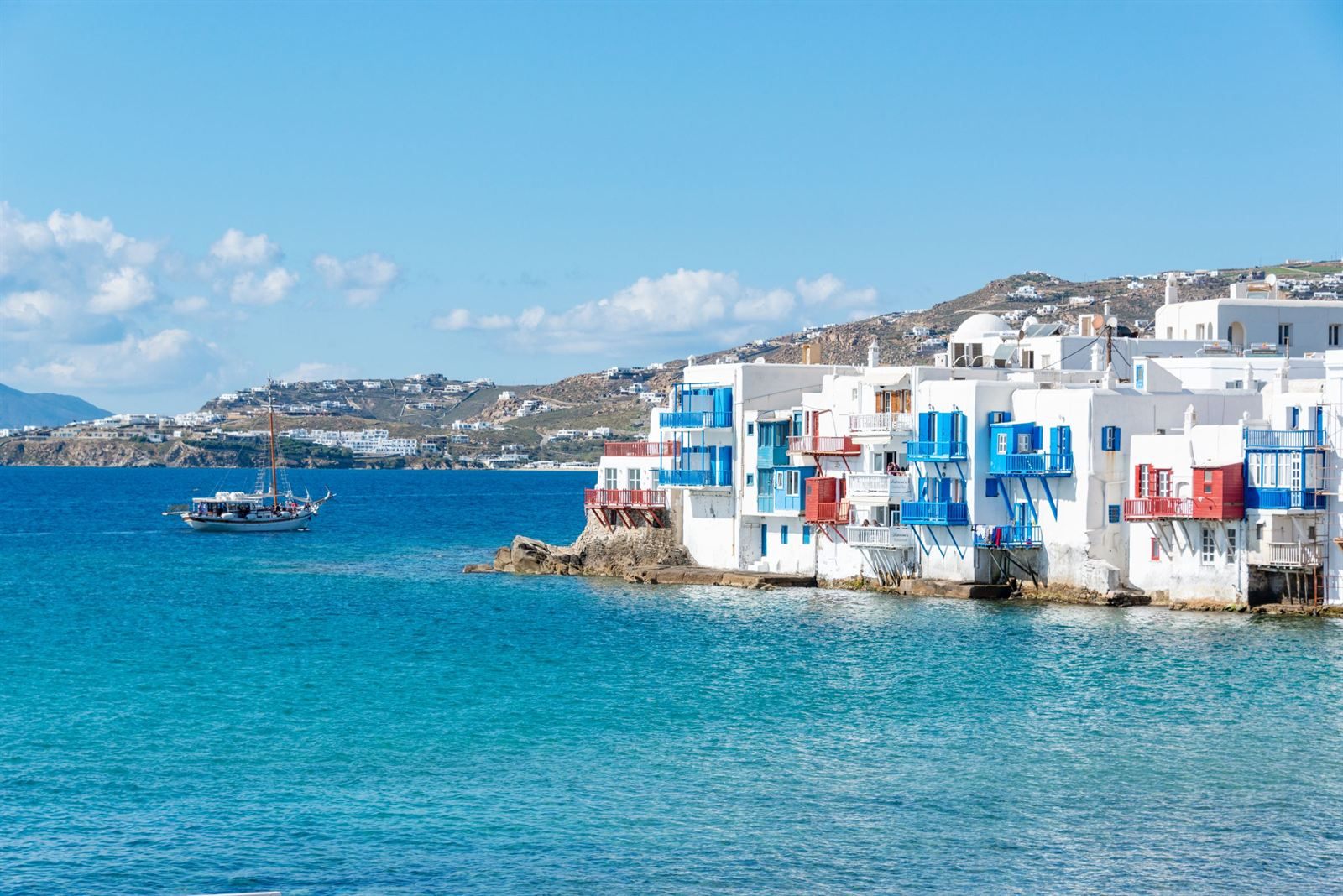 Whitewashed houses with colorful balconies over the water in Little Venice, Mykonos, with a sailboat offshore