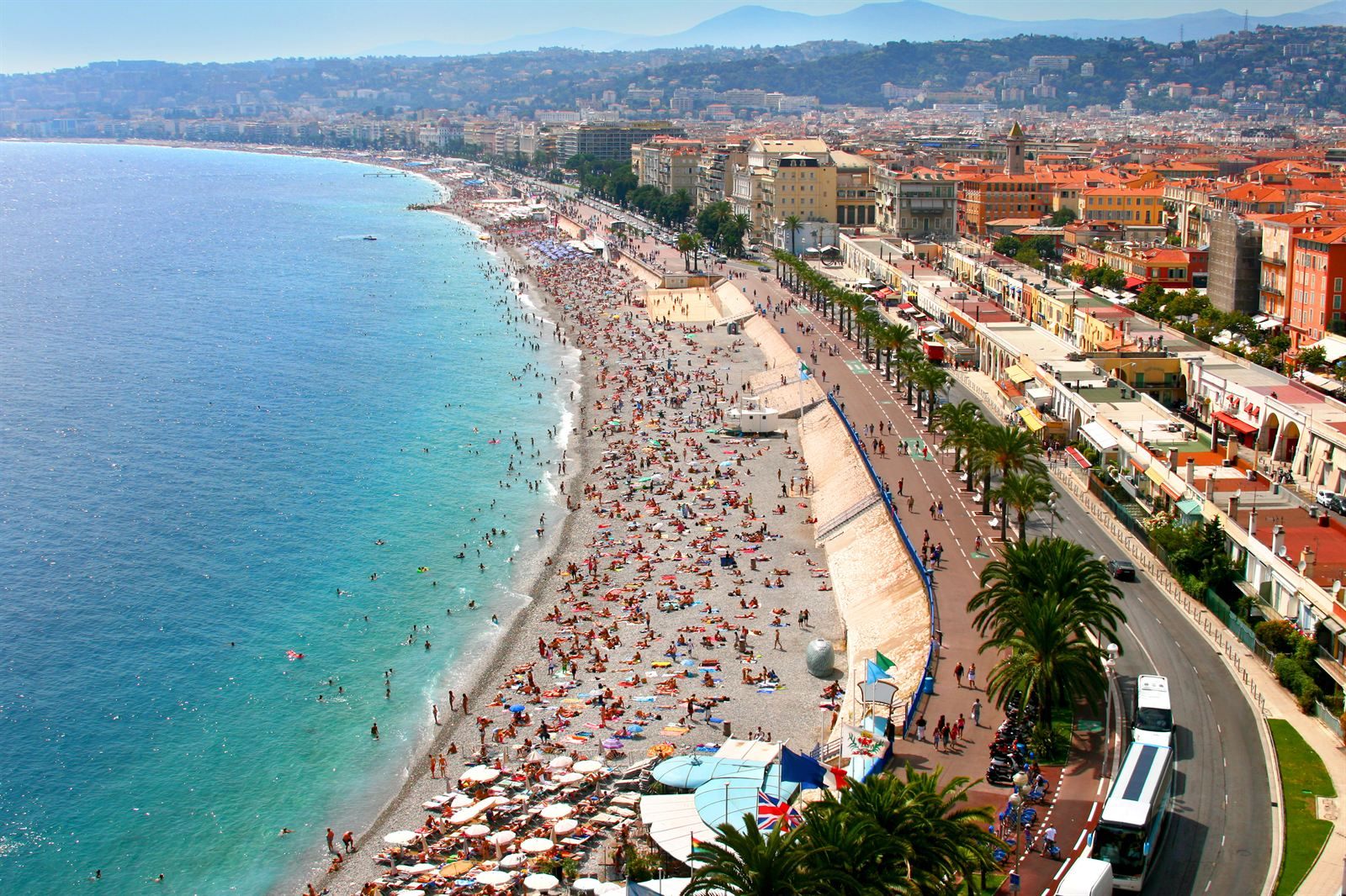 Crowded pebble beach and palm-lined Promenade des Anglais along the turquoise water in Nice