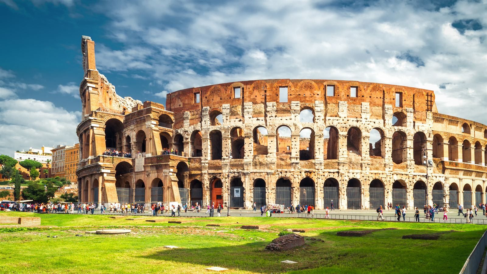 The Colosseum in Rome under a blue sky, with visitors on the lawn in front